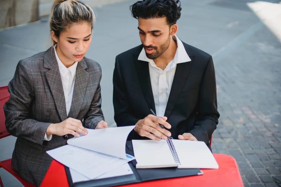 Two people in business suits sit at a small table looking over documents between them.